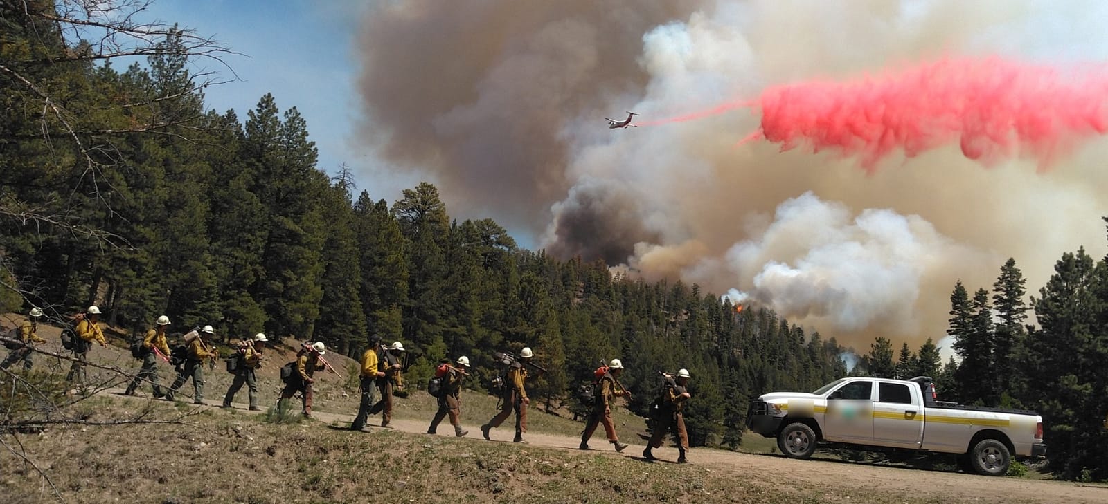 A wildland fire crew walks a dirt road while an air tanker drops retardant over a smoking ridge.